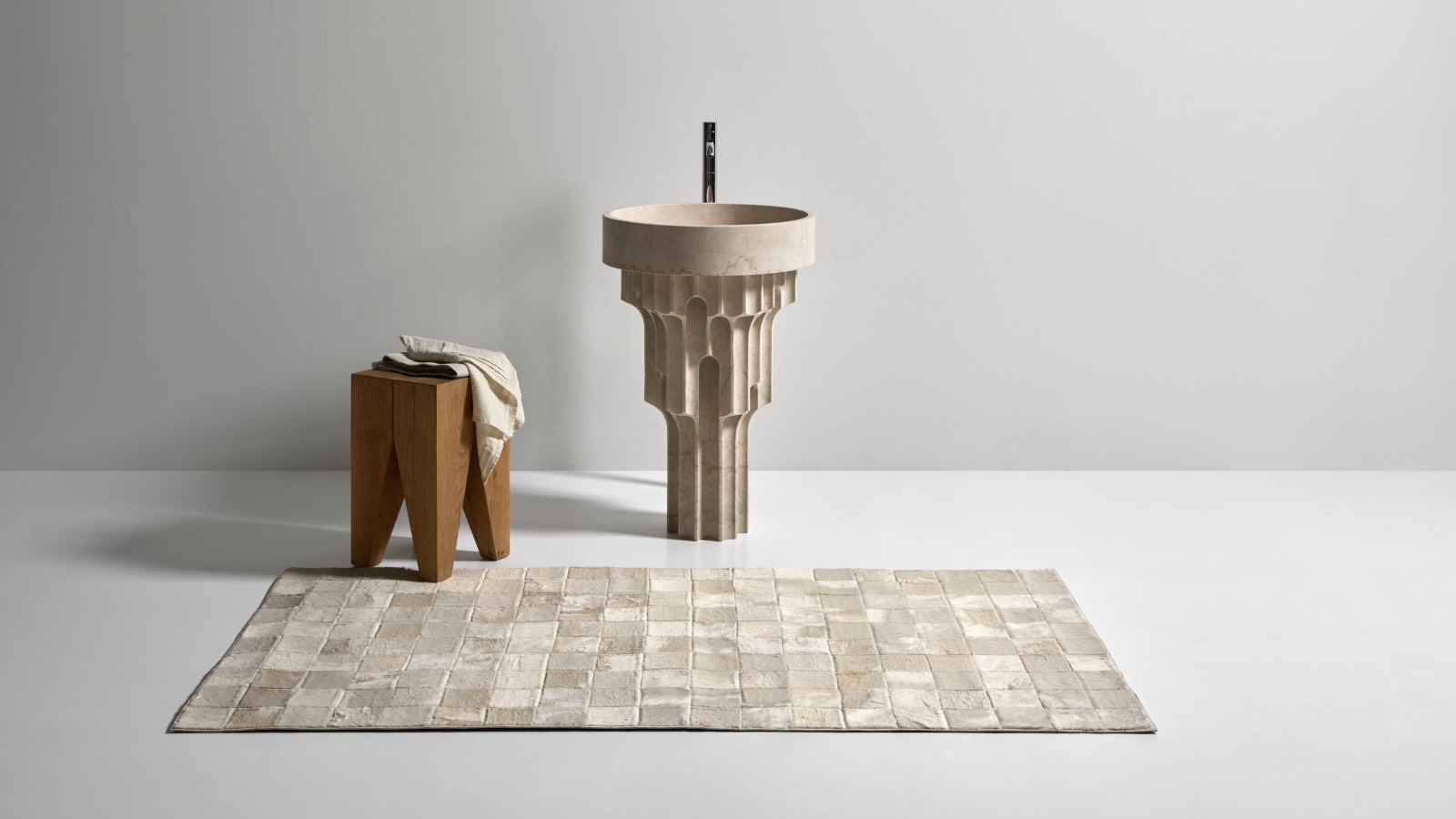 A sculptural stone pedestal sink with an arched design stands beside a geometric wooden stool and a rectangular textured rug on a minimalist white floor.