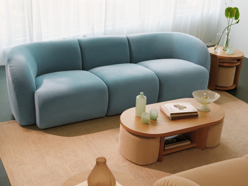 A modern living room with a blue curved sofa, oval wooden coffee table with books, glassware, and two round side tables, set on a beige rug near large windows.