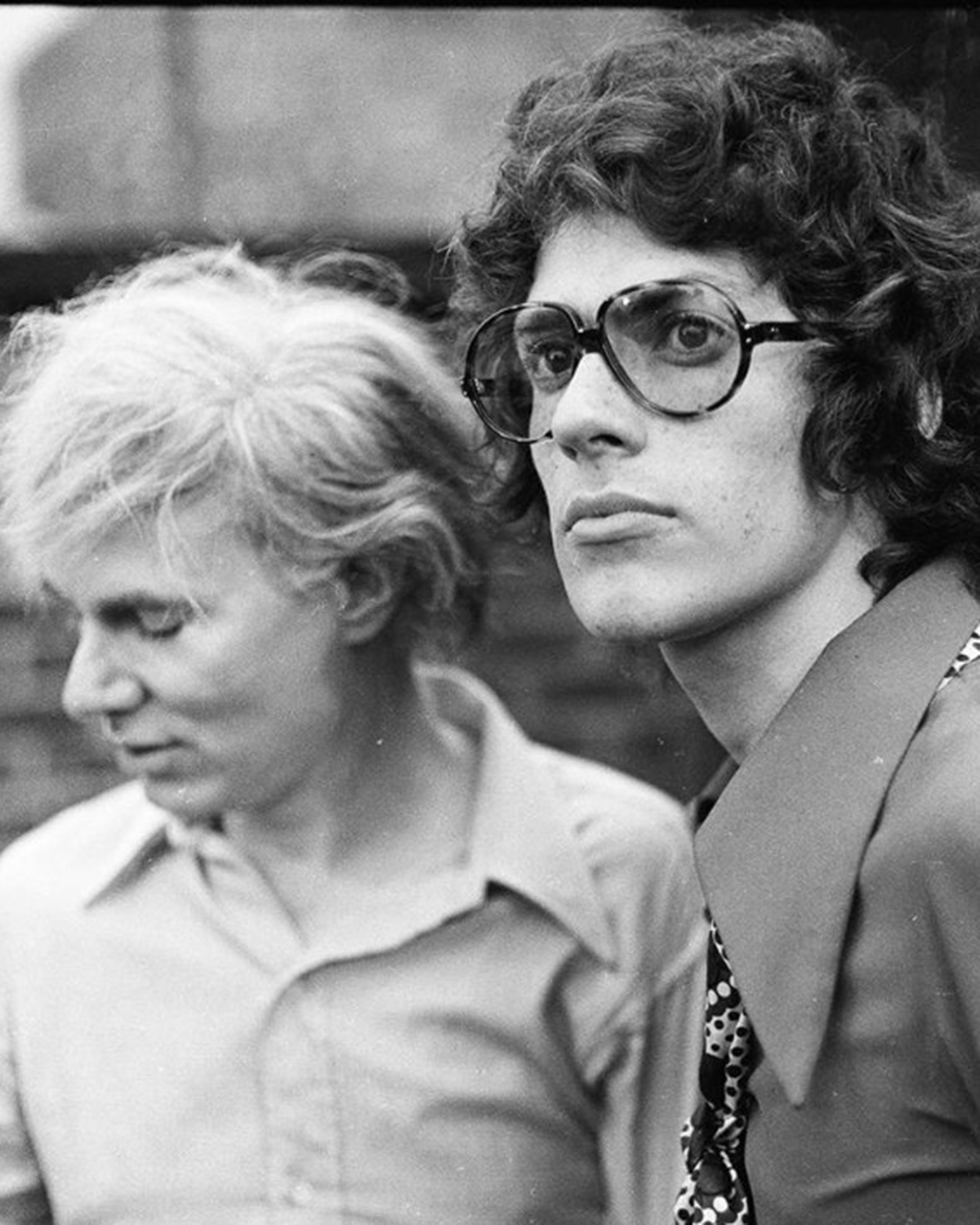 Two people in collared shirts, one with large glasses looking at the camera and the other looking down, appear in a black-and-white photo, evoking the timeless atmosphere of past July events.