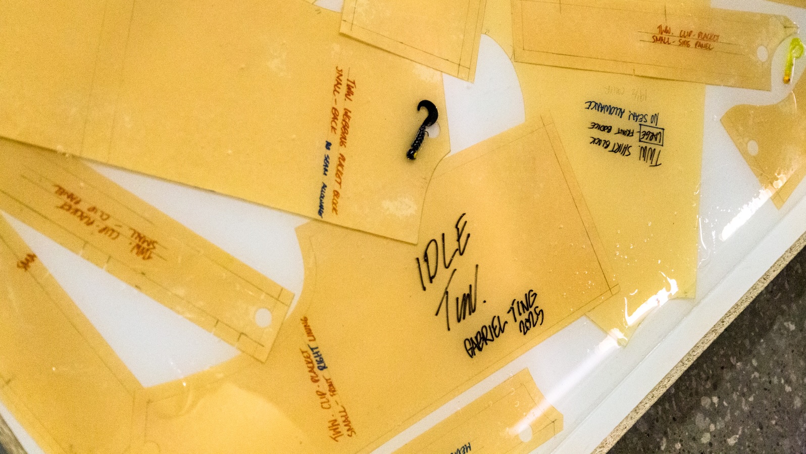 Several translucent garment pattern pieces with handwritten labels are displayed on a white surface; one reads “IDLE TWIN.” A small black hook is resting among the patterns.