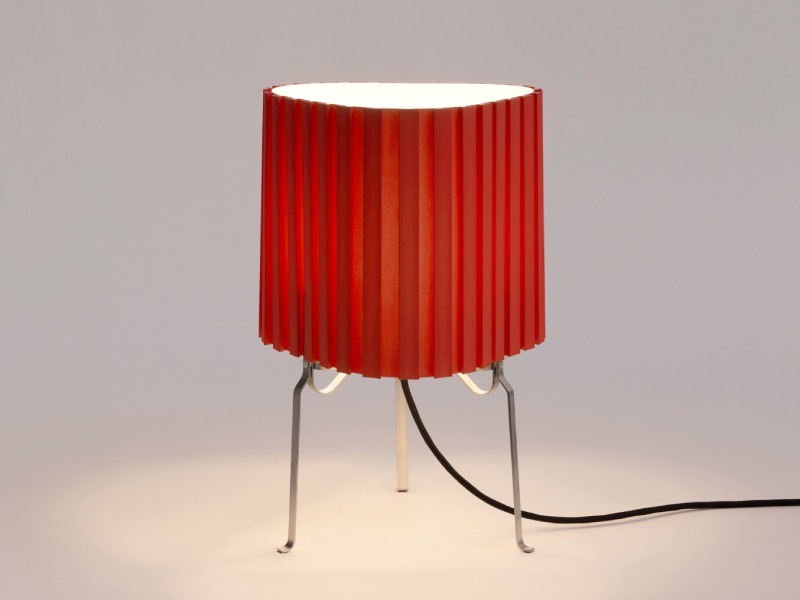 A small table lamp with a red pleated lampshade and three thin metal legs is shown on a plain surface with its power cord visible.