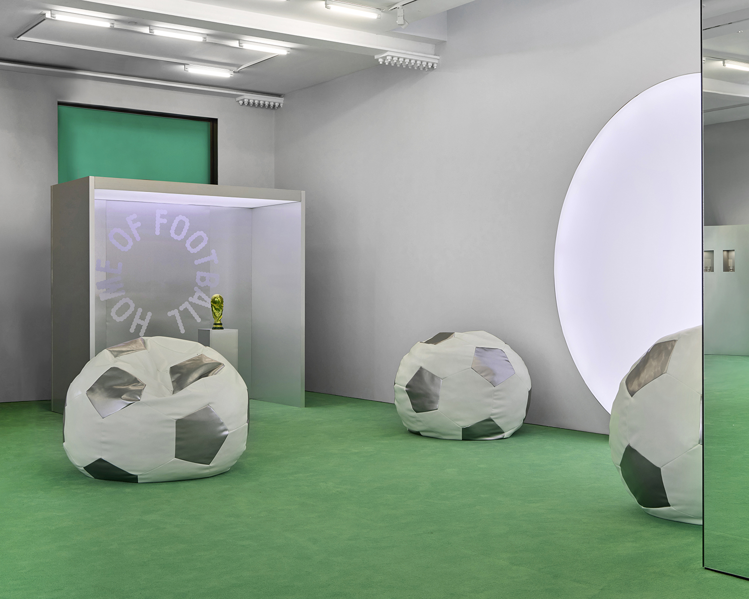 A modern room with a green floor, soccer ball-shaped bean bags, a display case containing a trophy, and a circular light feature on the wall—perfect for hosting July events or celebrating game day.