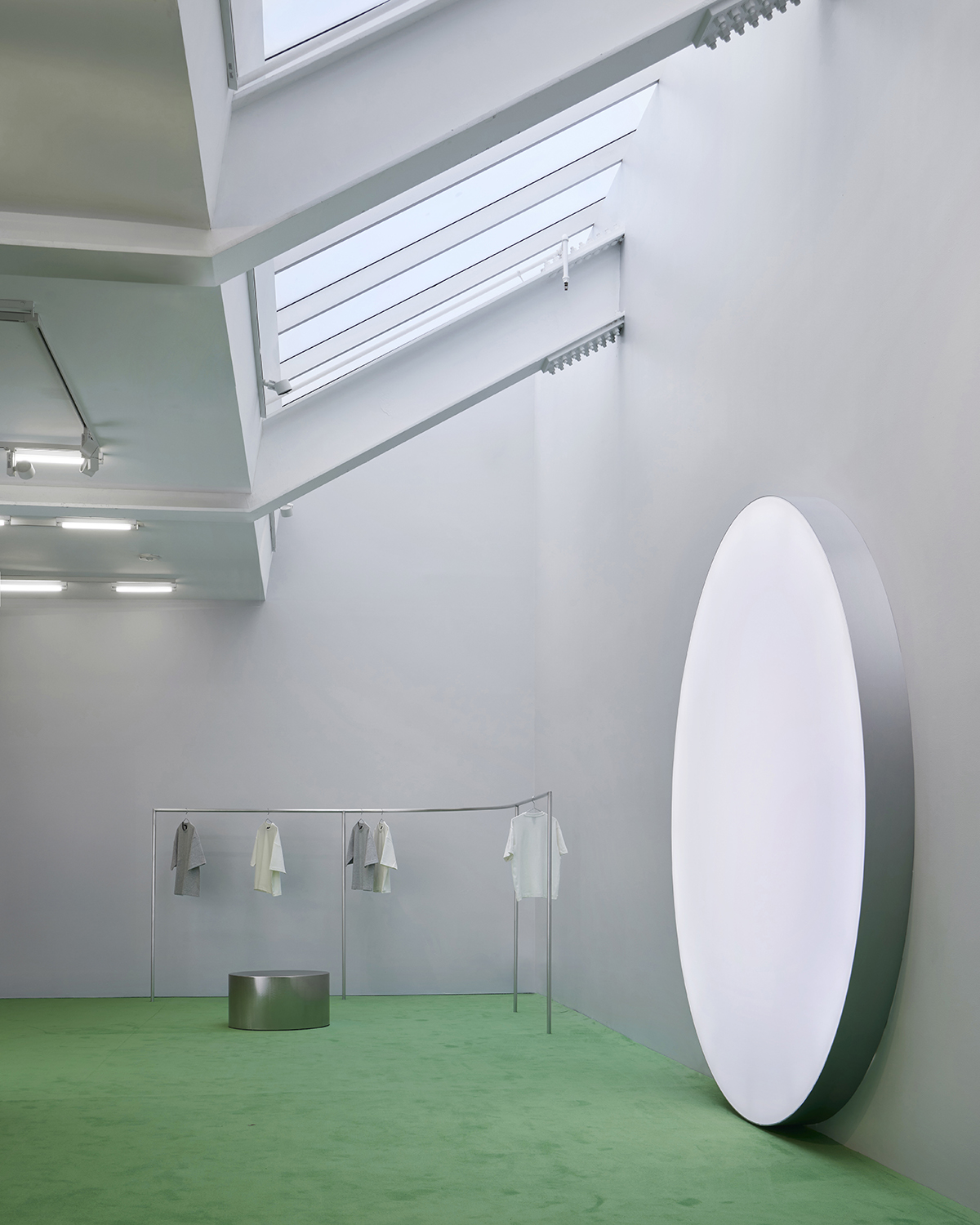Minimalist gallery space with green carpet, skylight, large illuminated oval wall fixture, circular pedestal, and clothing displayed on thin racks—perfect for showcasing July events in a contemporary setting.