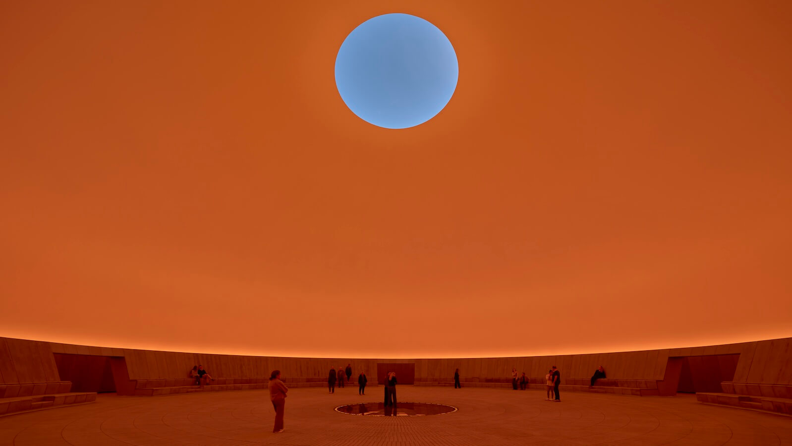 People stand inside a large circular room with orange-toned walls and a round opening in the ceiling, revealing a bright blue sky.