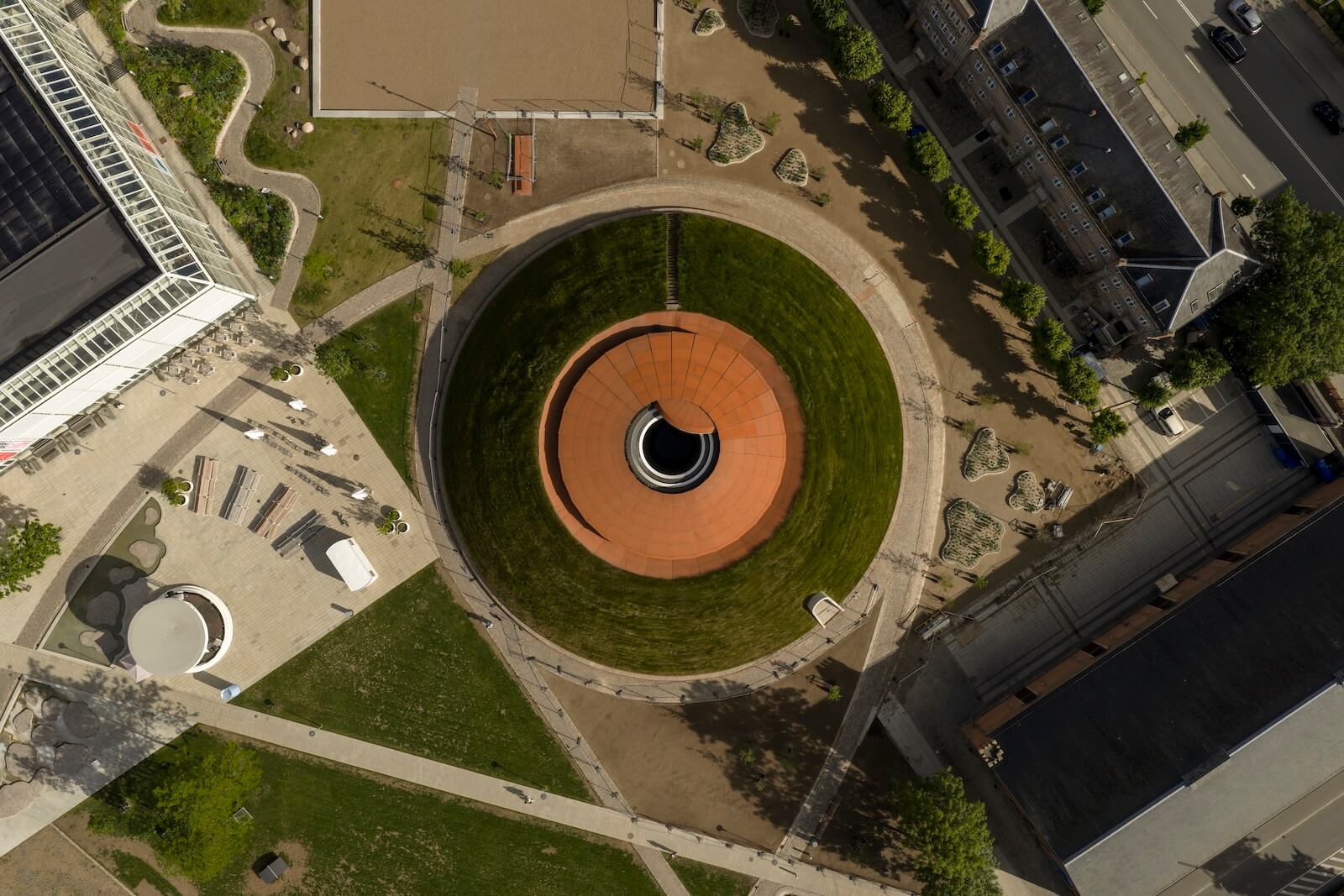 Aerial view of a circular, sunken grass area with a central red structure, surrounded by walkways, buildings, and green spaces.