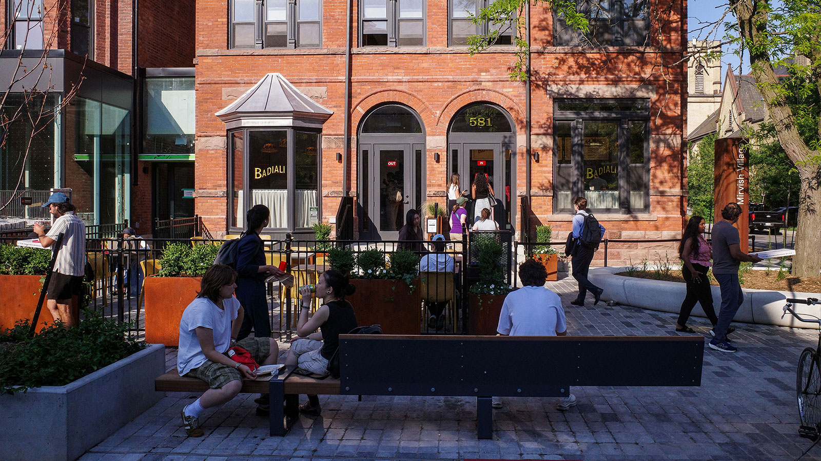 People sit and walk outside a red-brick building with large windows and a patio area on a sunny day.