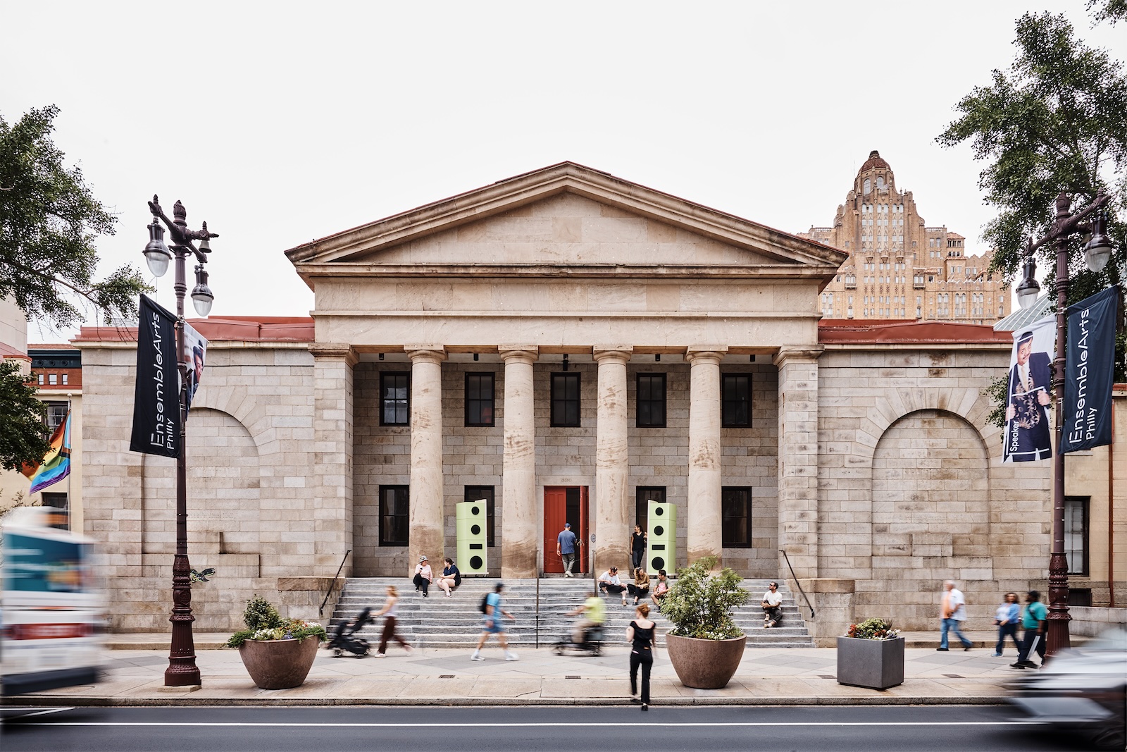 A neoclassical building with six columns and stone steps at the entrance, surrounded by people walking on the sidewalk and banners hanging from streetlights.