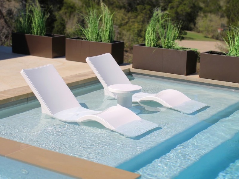 Two white lounge chairs and a small round table sit partially submerged in a shallow pool, with green plants in brown planters along the edge.