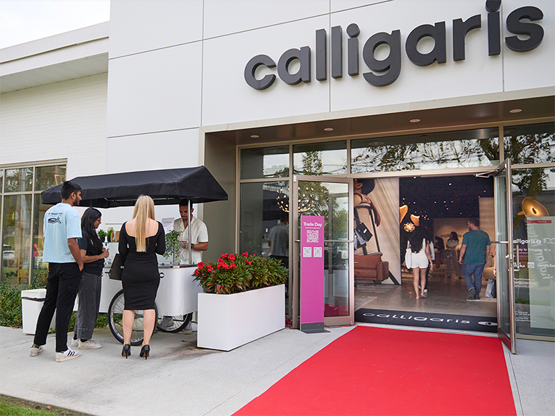 Four people stand by a cart outside the entrance of a Calligaris store, gathered for Tycos Drive Trade Day, with a red carpet leading inside and a large store sign above the door.