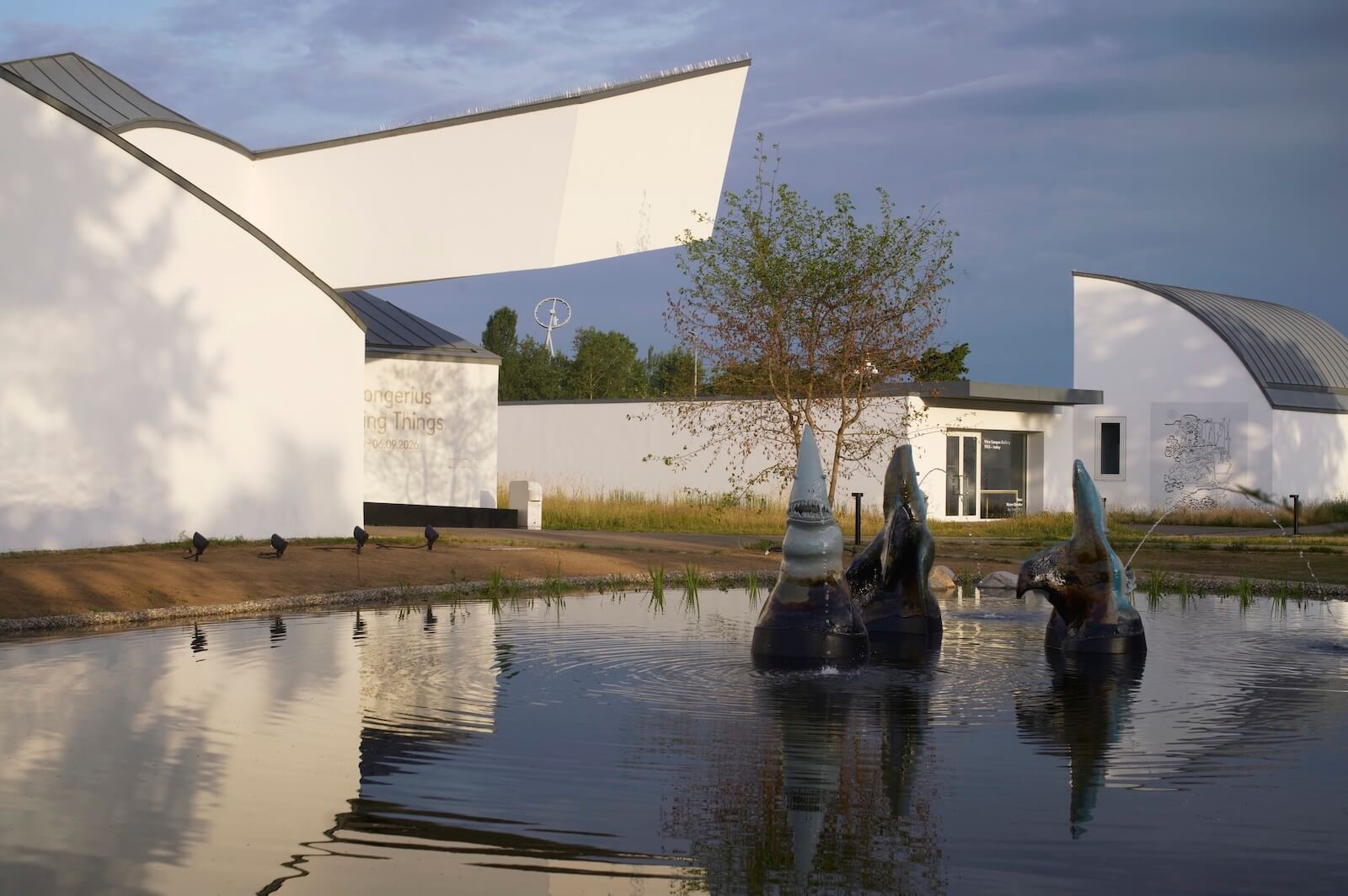 Modern white buildings with curved roofs stand near a pond with abstract sculptures and a small tree in the foreground, reflecting in the water under a partly cloudy sky.