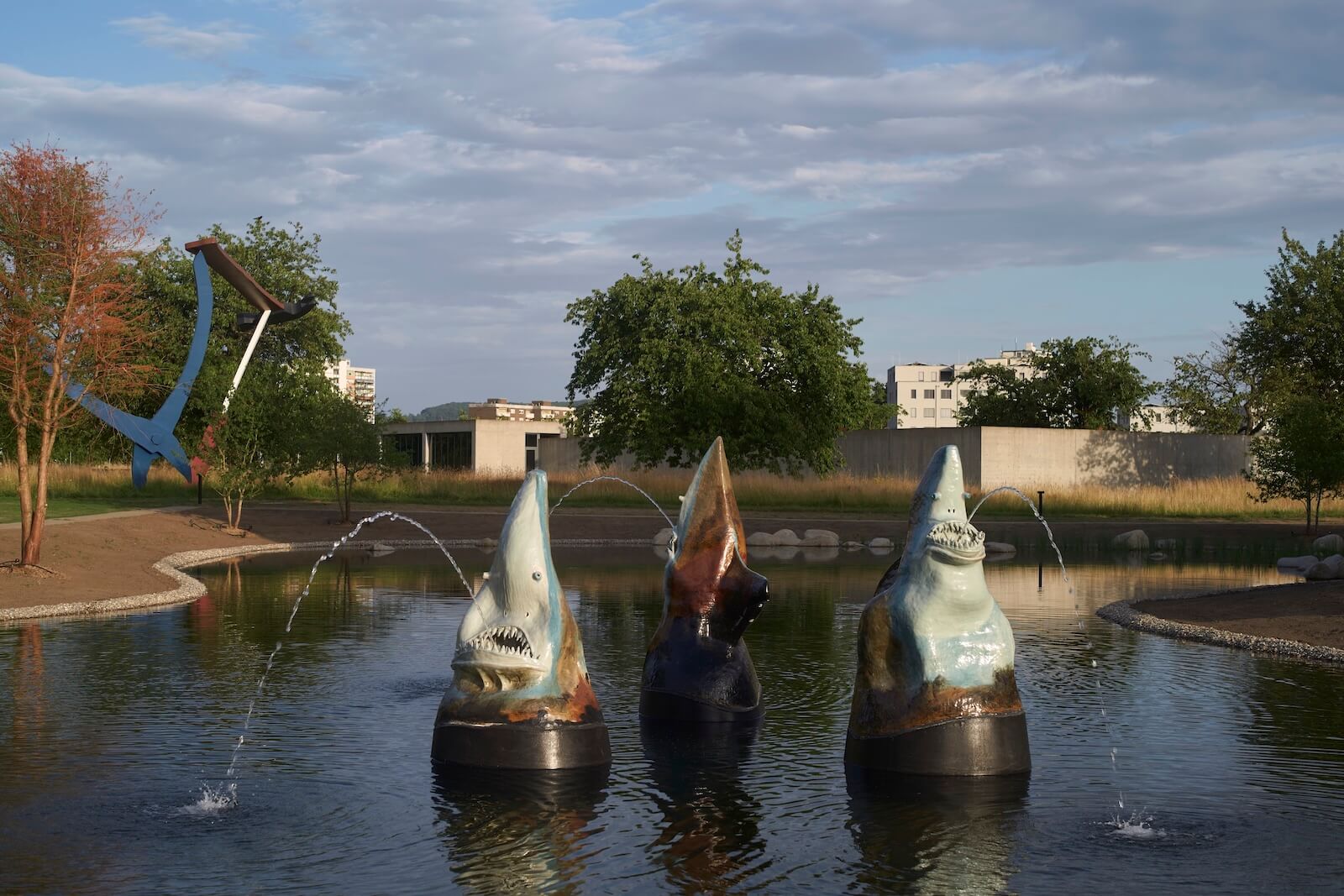 Three shark sculptures with open mouths spray water in a pond, with trees, grass, and buildings visible in the background under a partly cloudy sky.