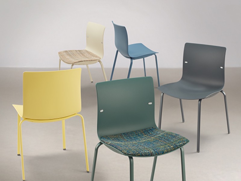 Five modern chairs with simple designs are spaced out on a neutral background. The chairs are yellow, beige, blue, dark grey, and green, with some seats made of fabric or woven material.