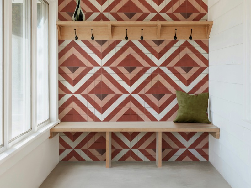 Wooden bench with a green pillow sits against a red and white geometric tile wall, beneath a shelf with hooks and a vase, next to large windows in a bright room.