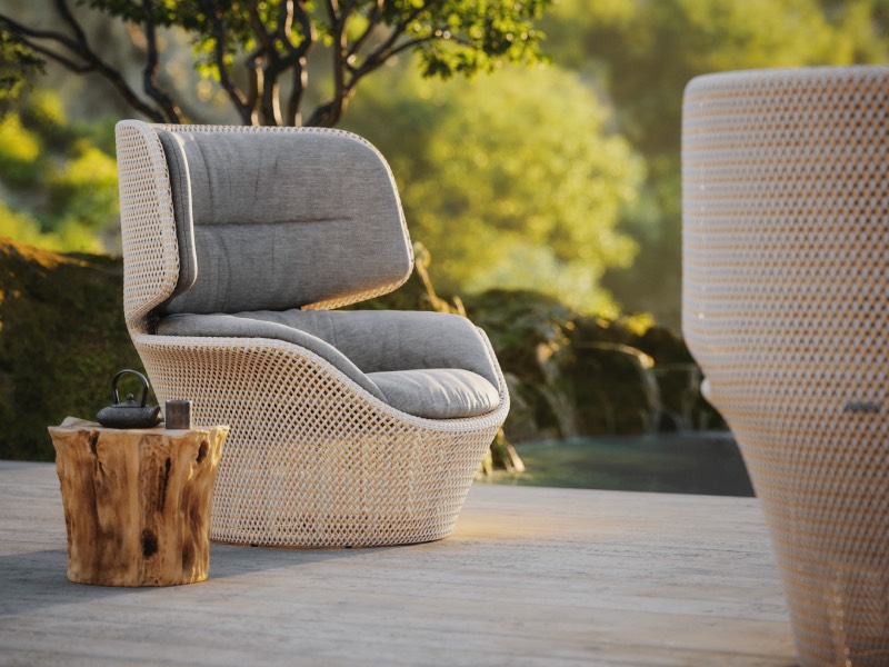 A wicker chair with gray cushions sits next to a small wooden table holding a teapot on a patio with greenery in the background.