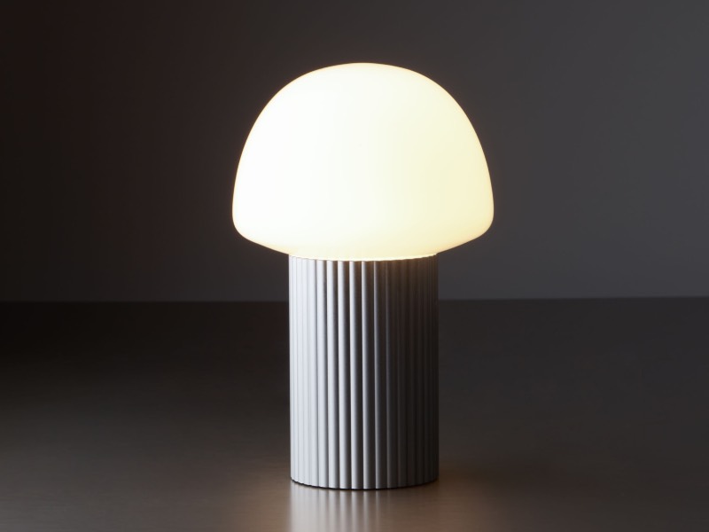 A table lamp with a ribbed cylindrical base and a rounded, glowing white lampshade sits on a smooth surface against a dark background.