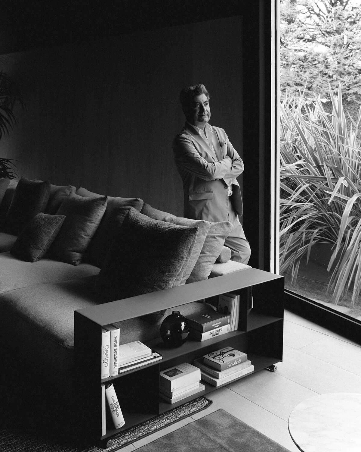 Antonio Citterio stands by a large window beside a sleek Groundpiece couch in a modern living room, with books and decor arranged on a low shelf and lush plants visible outside.
