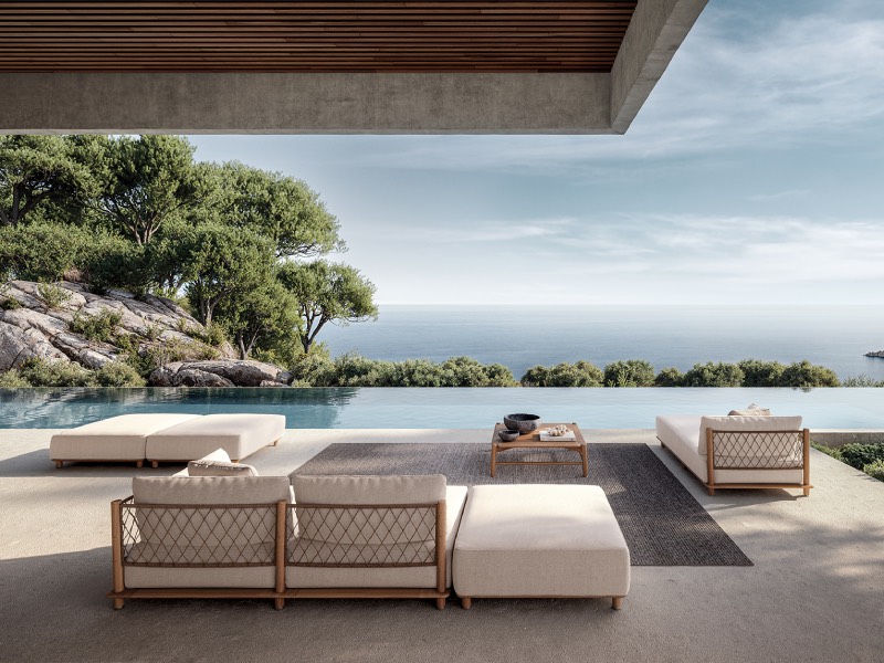 Modern outdoor patio with beige sofas and a coffee table overlooks an infinity pool, lush greenery, rocky landscape, and a calm sea under a partly cloudy sky.