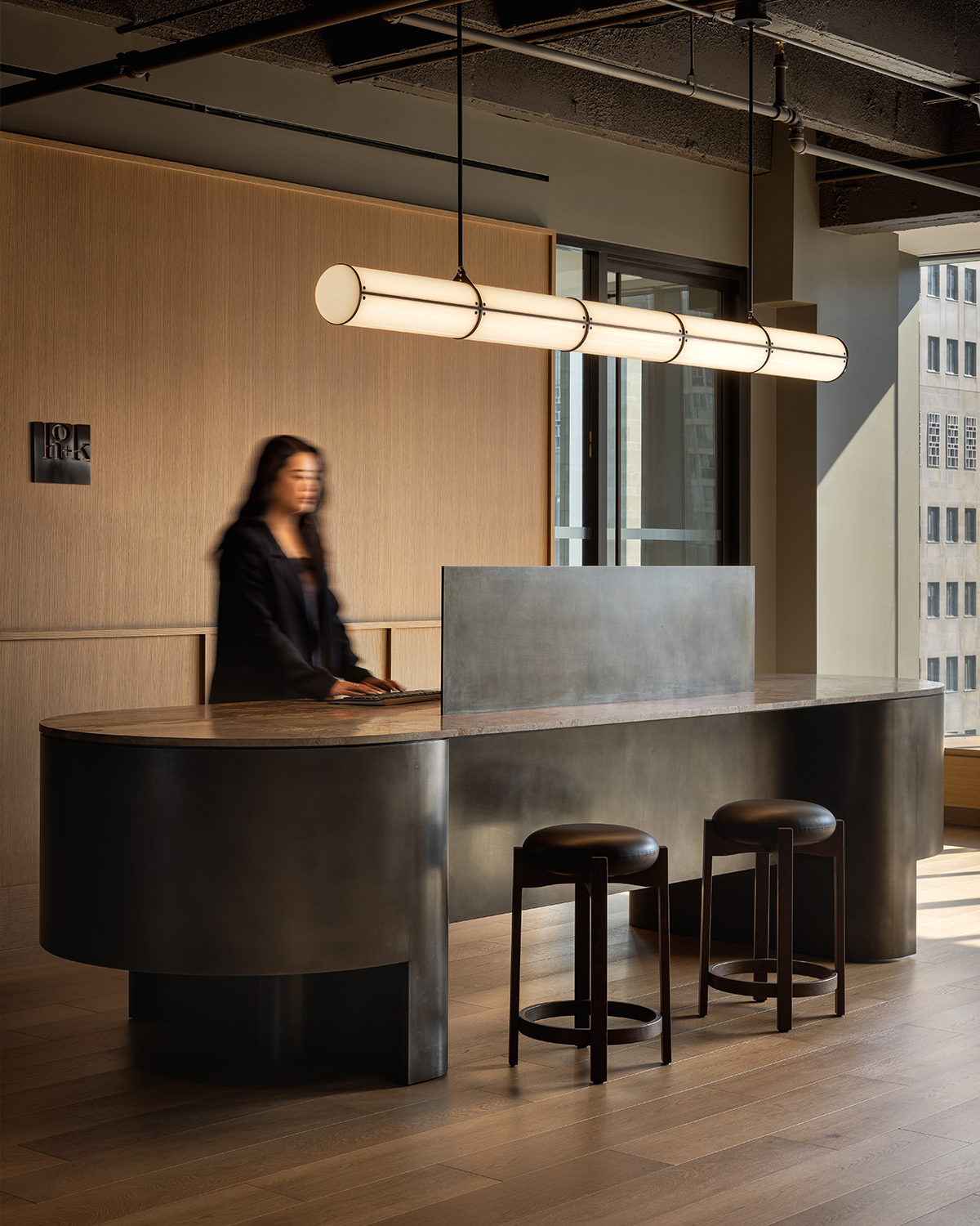 A woman stands at a curved black reception desk with two stools in HOK's Toronto office space, under a linear ceiling light.