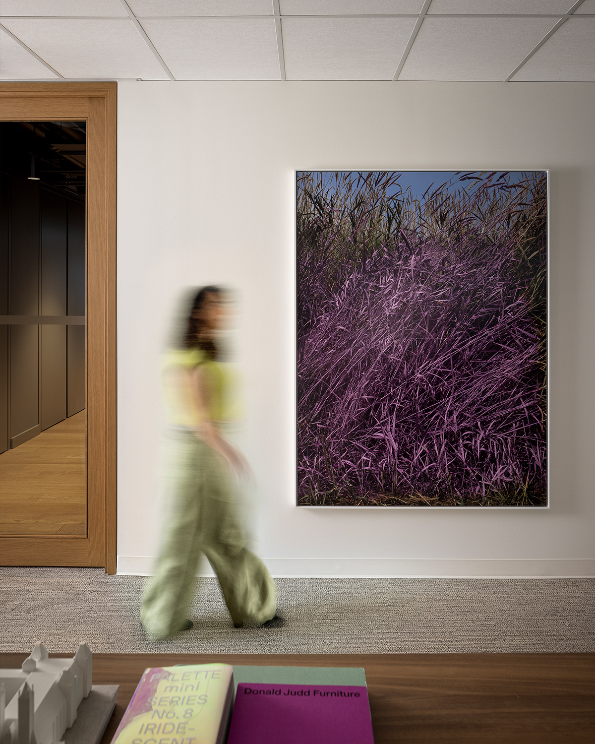 A blurred woman walks past a large framed photograph of purple grass in an office setting; books and a small architectural model sit on a nearby table.