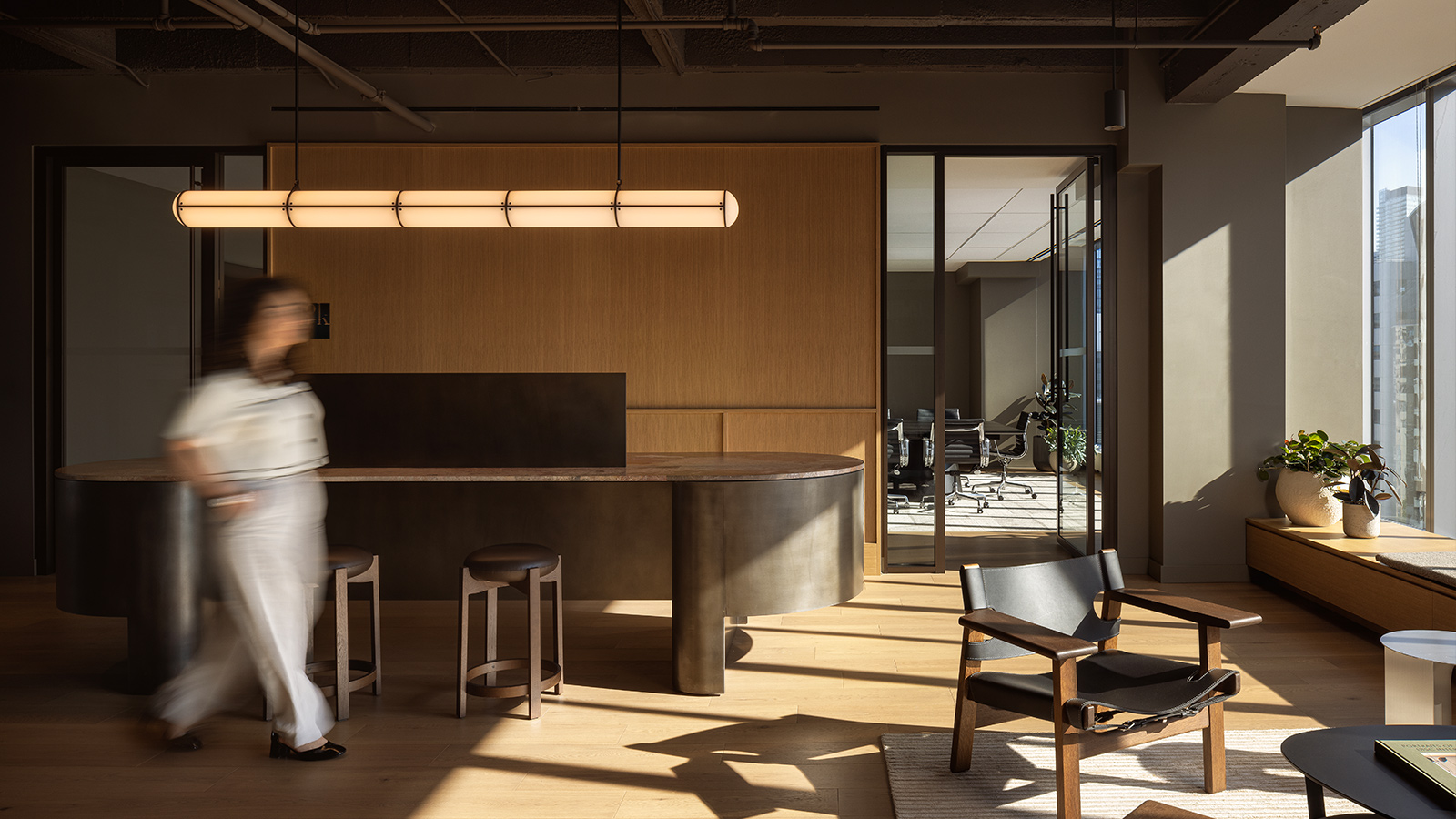 Modern office space with wooden floors, a curved black counter, stools, a meeting room in the background, a blurred person walking, and sunlight streaming through large windows.