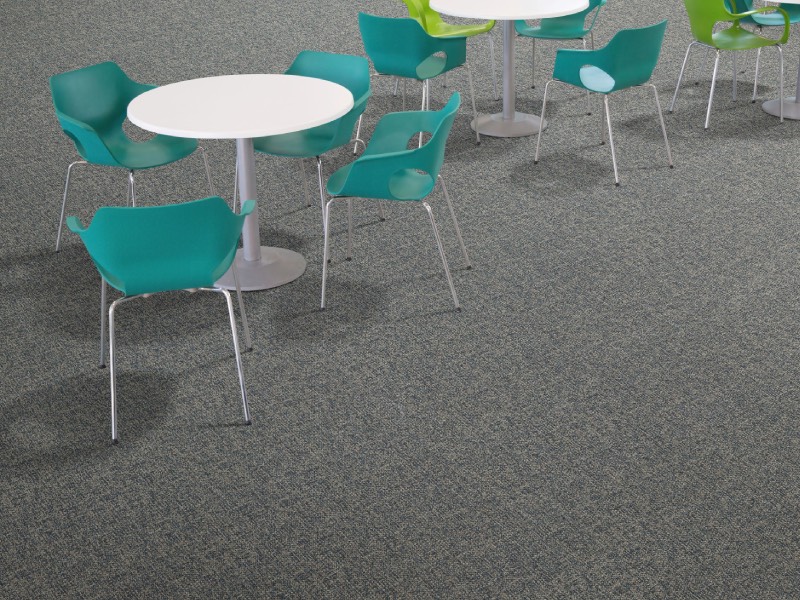 Several round white tables with green and blue plastic chairs are arranged on a gray carpeted floor in a modern, empty seating area.