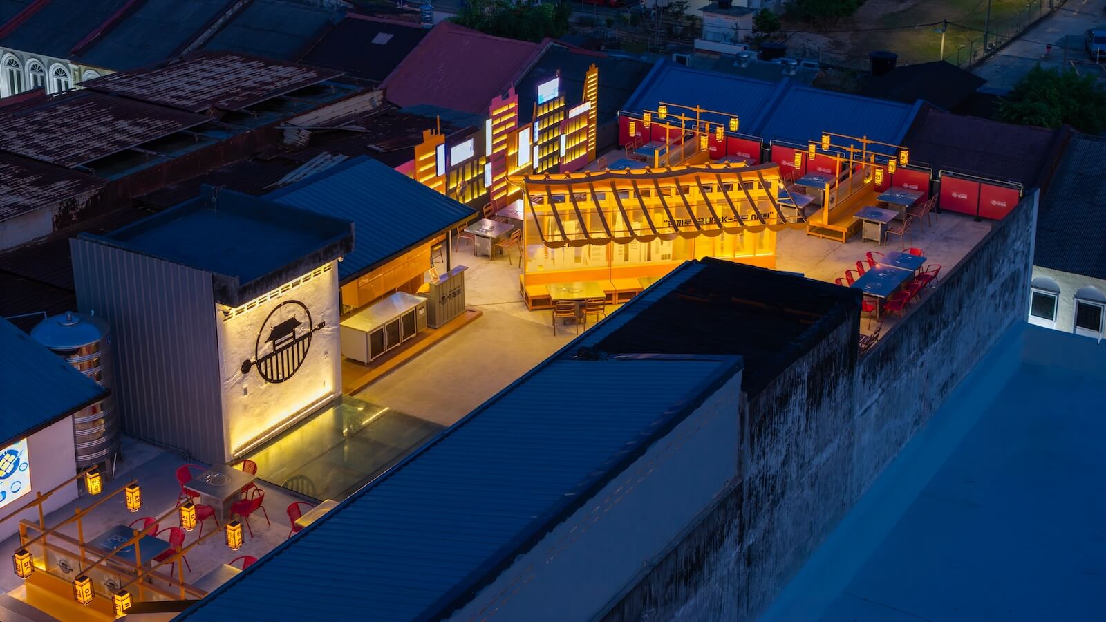 A rooftop restaurant with yellow lighting, red chairs, and multiple seating areas at night, surrounded by older buildings and rooftops.