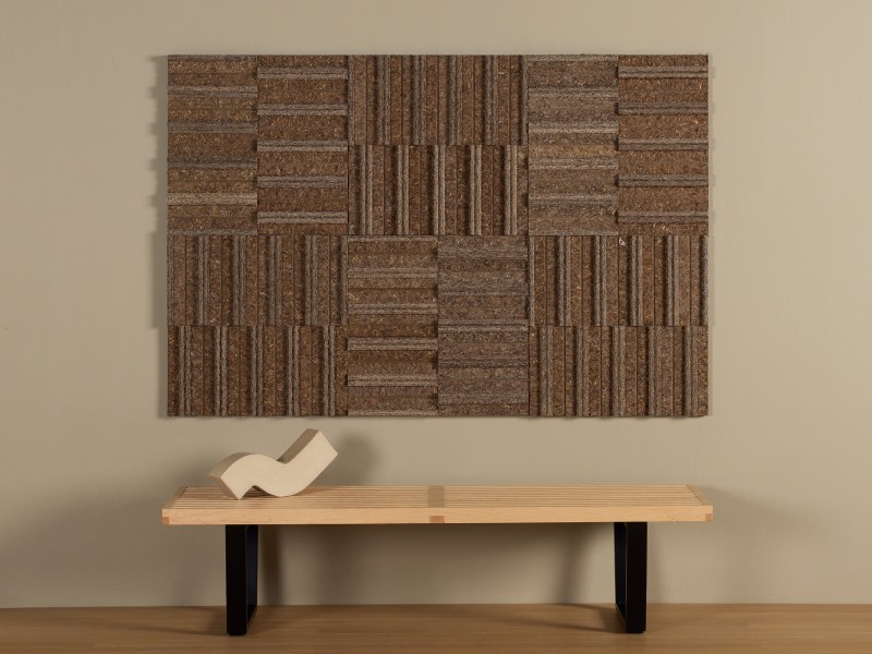 A wooden bench with black legs sits beneath a wall art piece made of textured, rectangular brown panels arranged in a geometric pattern.