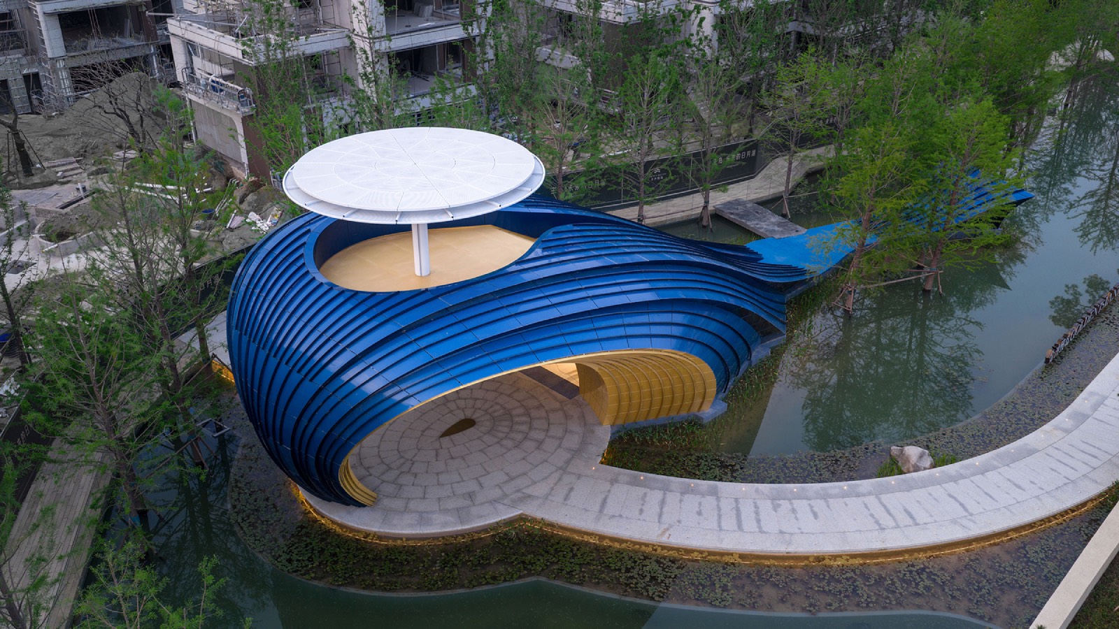 A blue, shell-shaped building with a white circular roof sits by a pond, connected to a curving walkway amid trees and surrounding construction.