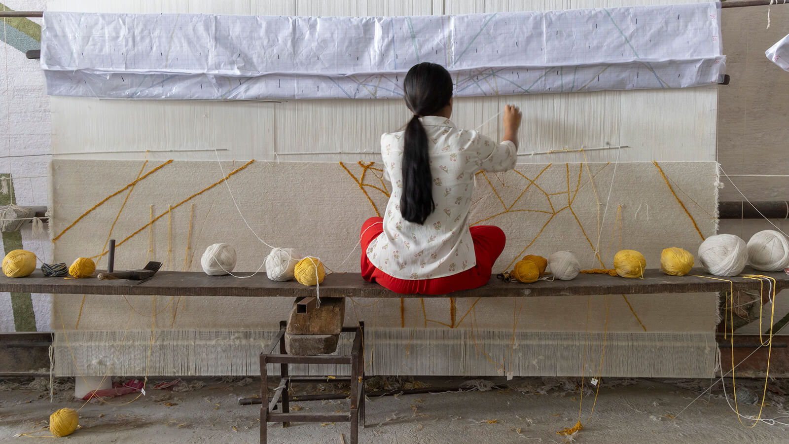 A person with long hair, in a white shirt and red pants, sits on a bench weaving on a large loom with white and yellow yarn in a workshop where zoometry and other creative matters come to life.