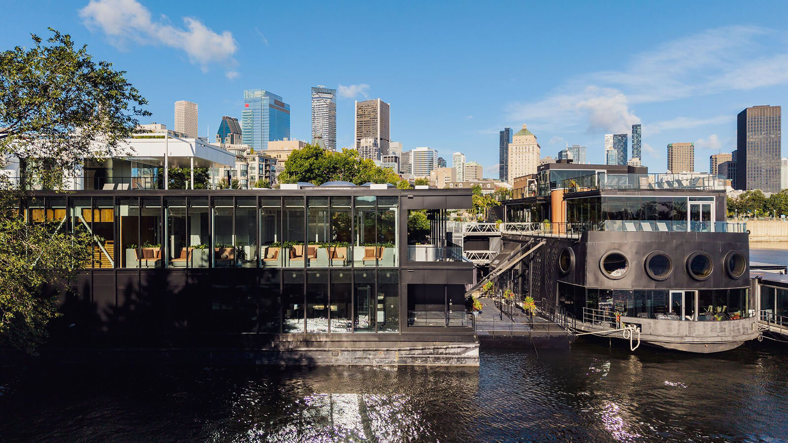 Modern black bota bota Pavillon on a waterfront with large windows, outdoor seating, and city skyscrapers visible in the background under a blue sky.