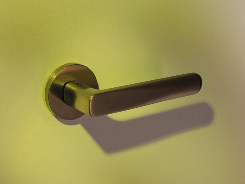 A metallic ROBOT6 door handle by Colombo Design America is mounted on a light-colored wall, casting a distinct shadow in soft lighting.