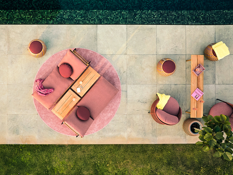Aerial view of an outdoor patio with pink TIBBO DEDON seating, round cushions, a wooden table, and stools near a pool. Some books and towels are placed on the furniture.