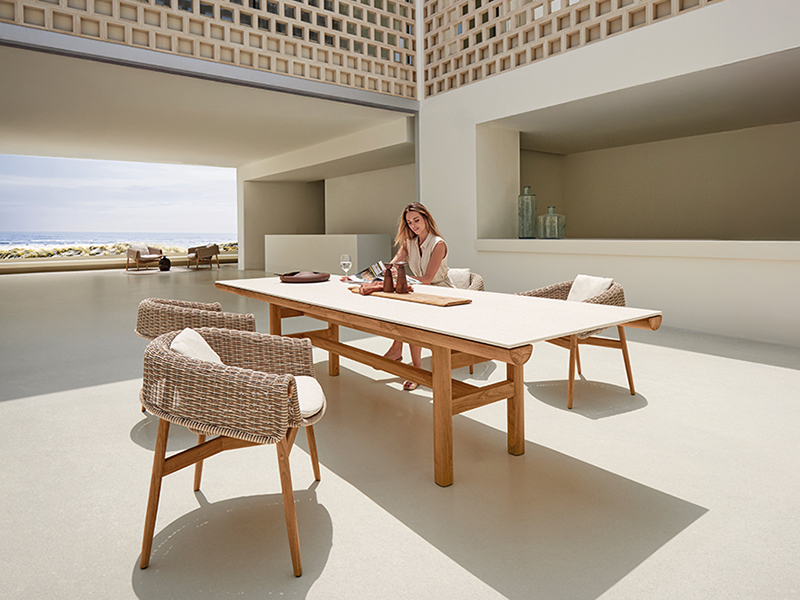 A woman sits alone at a large outdoor dining table with wicker chairs, writing in a notebook. The space is modern, open, and sunlit, featuring elegant rail gloster details alongside large windows and minimalistic decor.