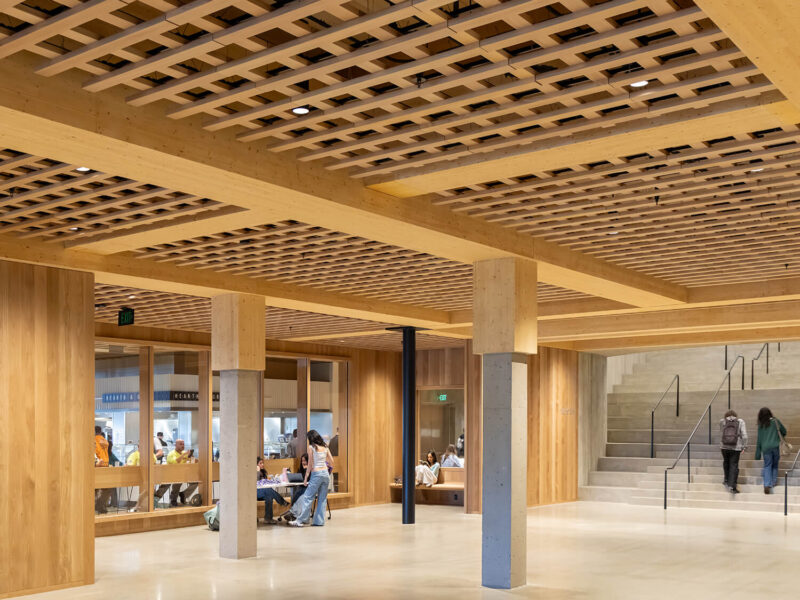 Spacious interior at Amherst College with wooden beams, a grid-patterned ceiling, glass walls, people sitting at tables, and others walking near a staircase.
