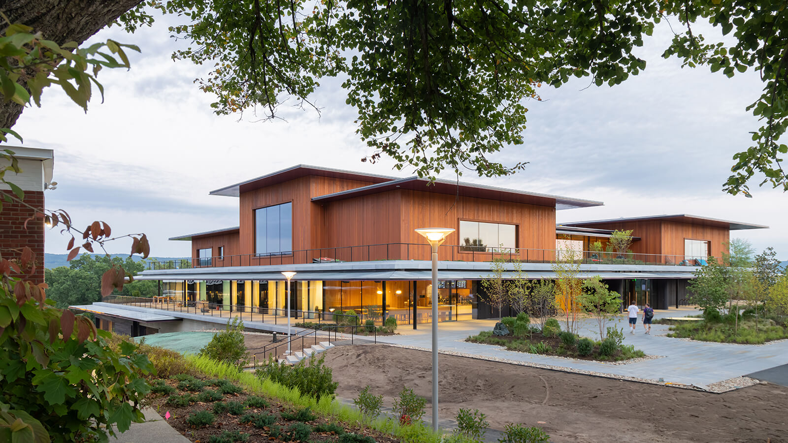 Modern two-story building at Amherst College with large windows and wood-paneled exterior, surrounded by landscaped greenery and a walkway, under a cloudy sky.
