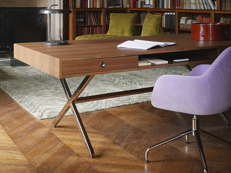 Modern wooden desk with an open drawer, papers, and a lamp, next to a luxurious Président Ligne Roset lavender swivel chair on a wooden floor, with bookshelves and green chairs in the background.