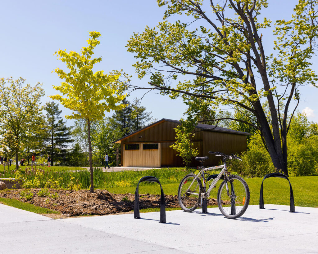 A black bicycle is parked at a bike rack on a paved path near a small wooden building, surrounded by green trees and grass under a clear sky.