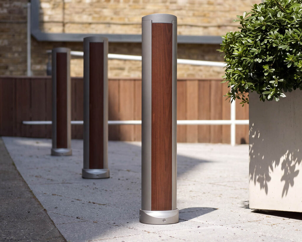 A row of modern, cylindrical bollards, each featuring sleek wood paneling, stands on a concrete path next to a large planter with green foliage.