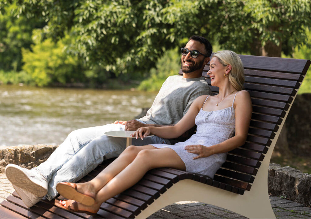 A man and woman sit together on a modern outdoor lounge chair by a river, smiling and enjoying a sunny day.