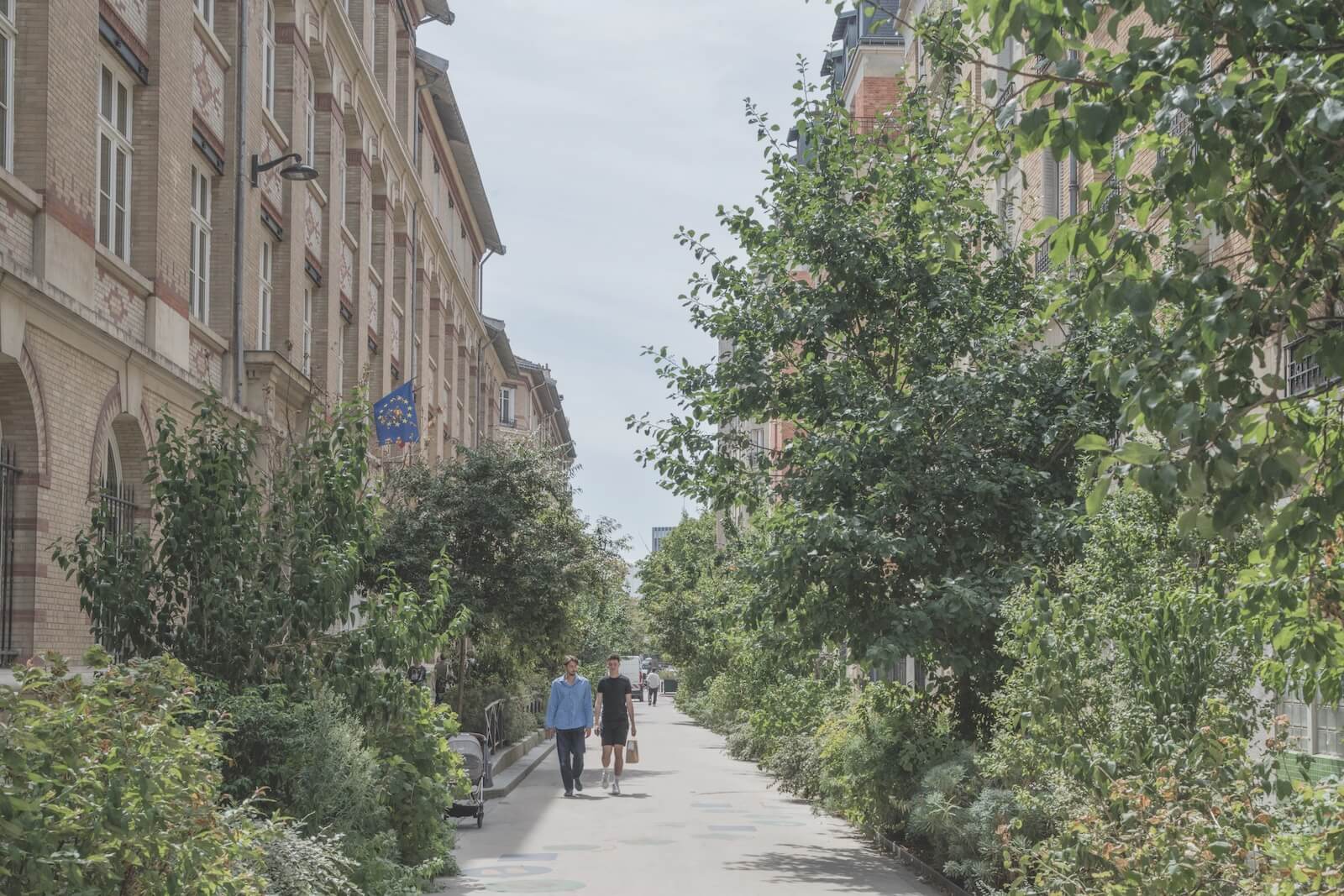 Two people walk down a tree-lined pedestrian path between brick buildings on a sunny day; an EU flag hangs from one building.