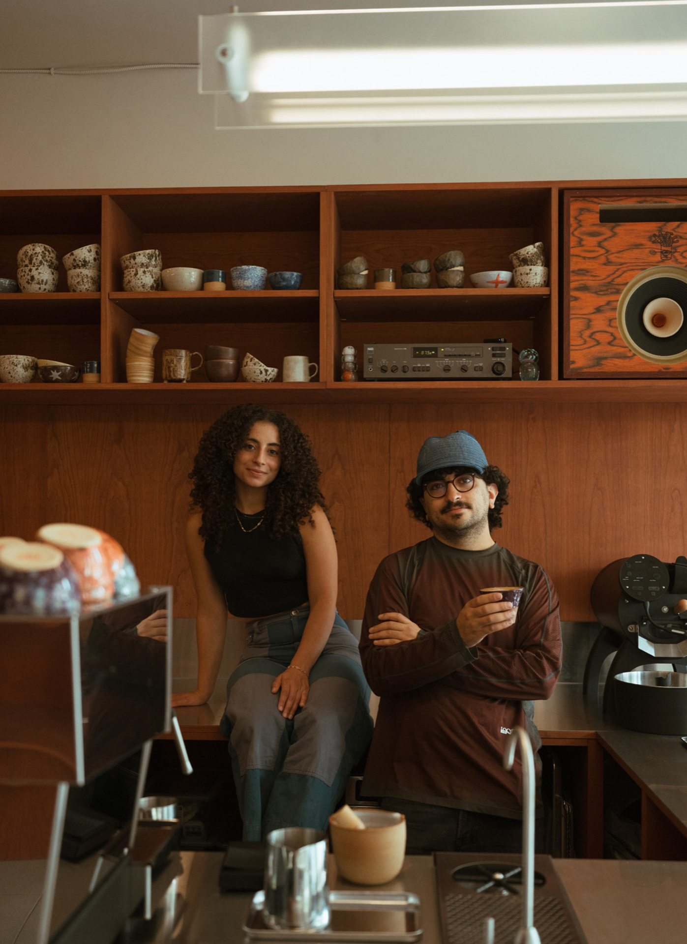 Two people pose in a coffee shop, surrounded by cups and equipment, with shelves of ceramics and a speaker behind them.