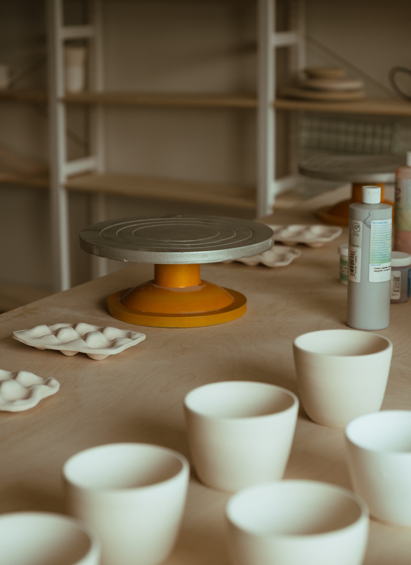 A pottery wheel sits on a wooden table with unglazed ceramic cups, paint bottles, and tools in a studio setting.
