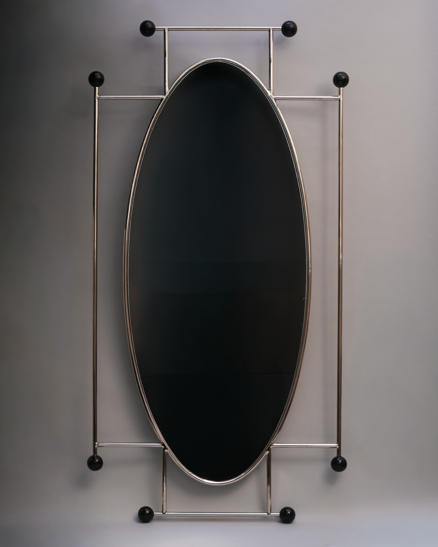A large, oval-shaped mirror with a metallic frame and black spherical accents at the corners, set against a plain gray background.