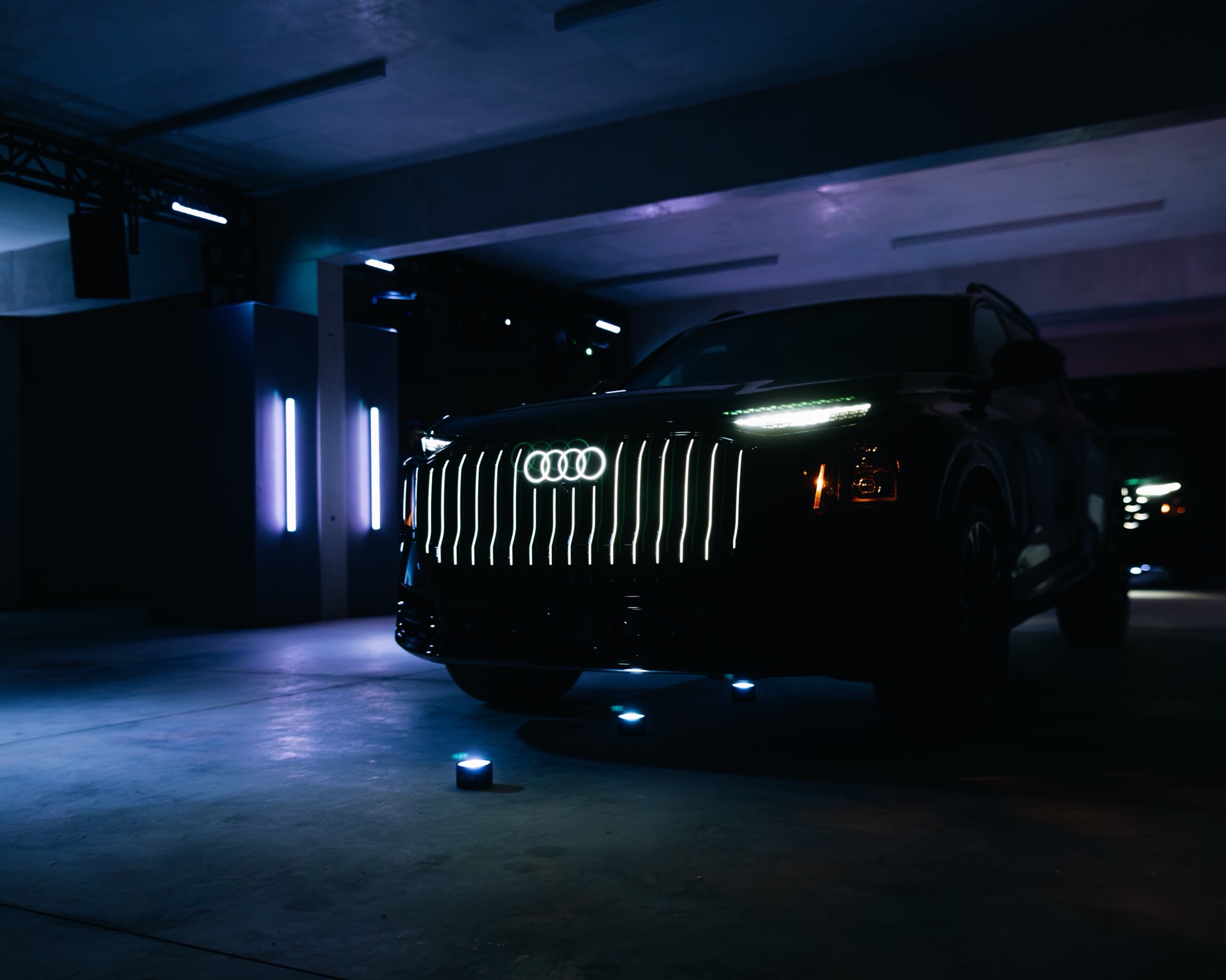 A dark Audi Q9 SUV with an illuminated front grille and logo, parked in a dimly lit space with vertical blue accent lights.