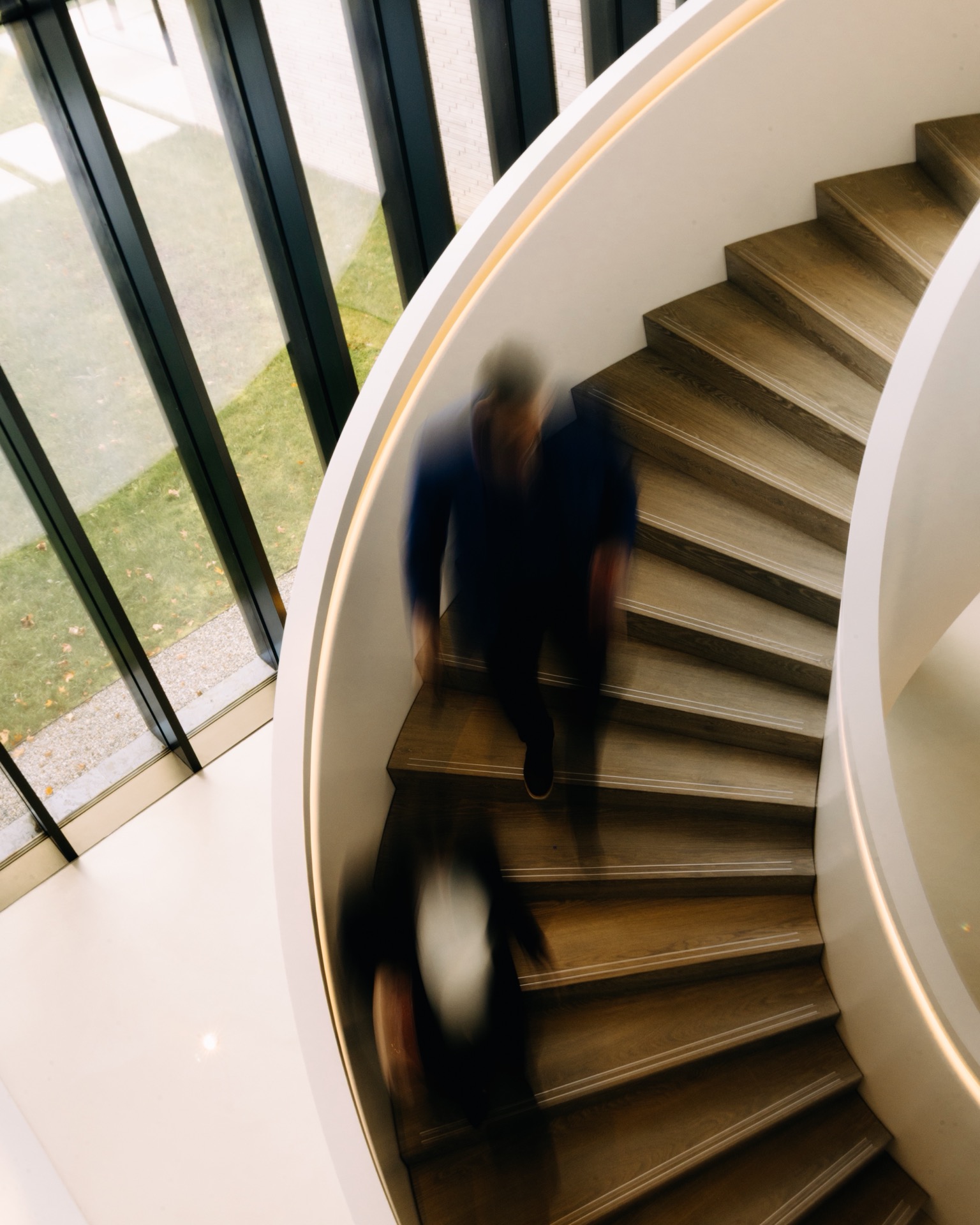A blurred figure walks down a curved wooden staircase beside floor-to-ceiling windows, evoking the sleek lines of an Audi Q9.