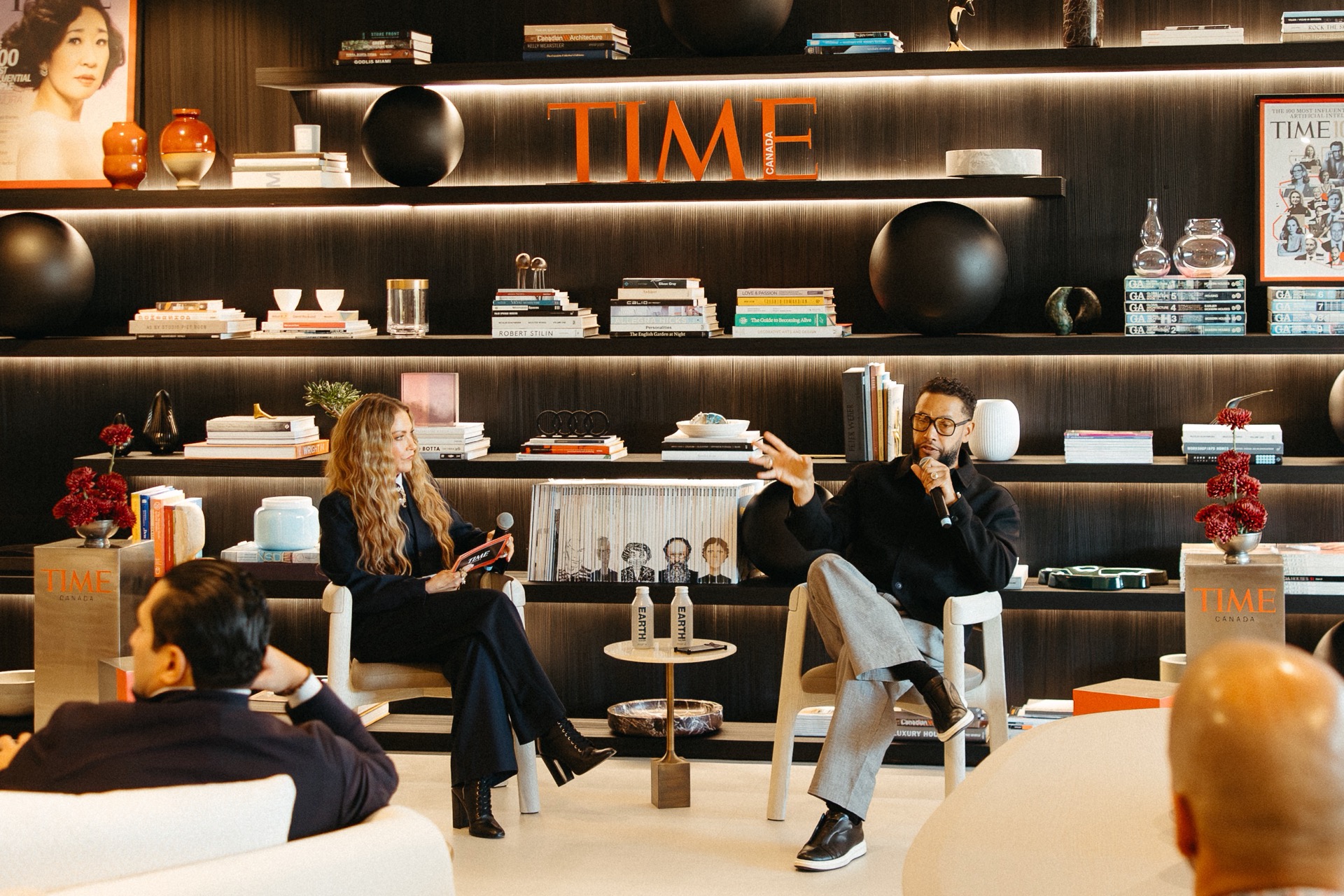 A woman and a man sit in armchairs on a stage in front of an Audi Q9 bookshelf, speaking into microphones during a panel discussion.