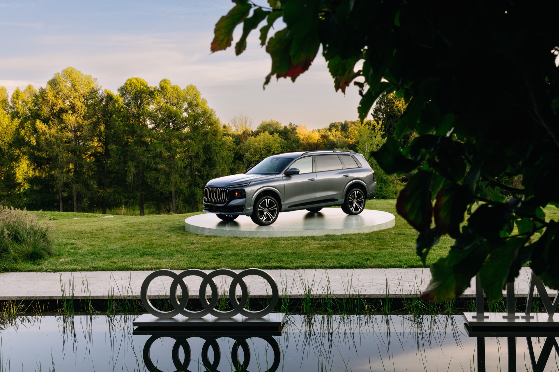 A grey Audi Q9 on a circular display platform in a grassy field, with an Audi logo sculpture in the foreground pond.