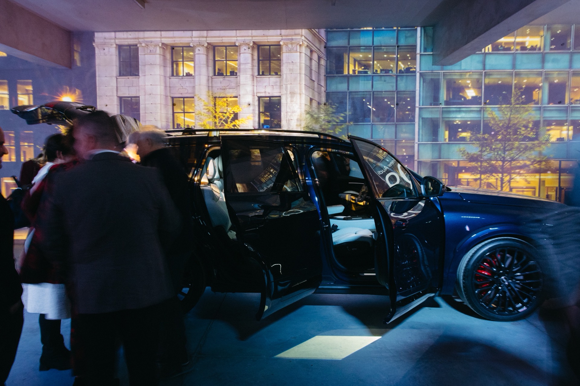 People standing around a dark blue Audi Q9 with its doors open in an urban setting.