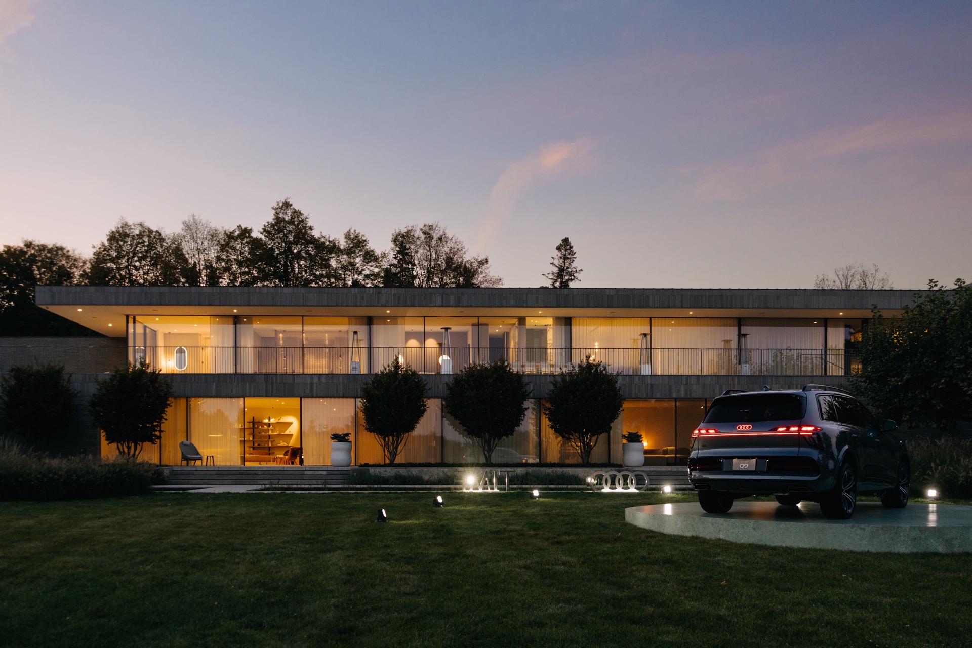 An Audi Q9 parked on a platform in front of a modern, two-story glass house at dusk.