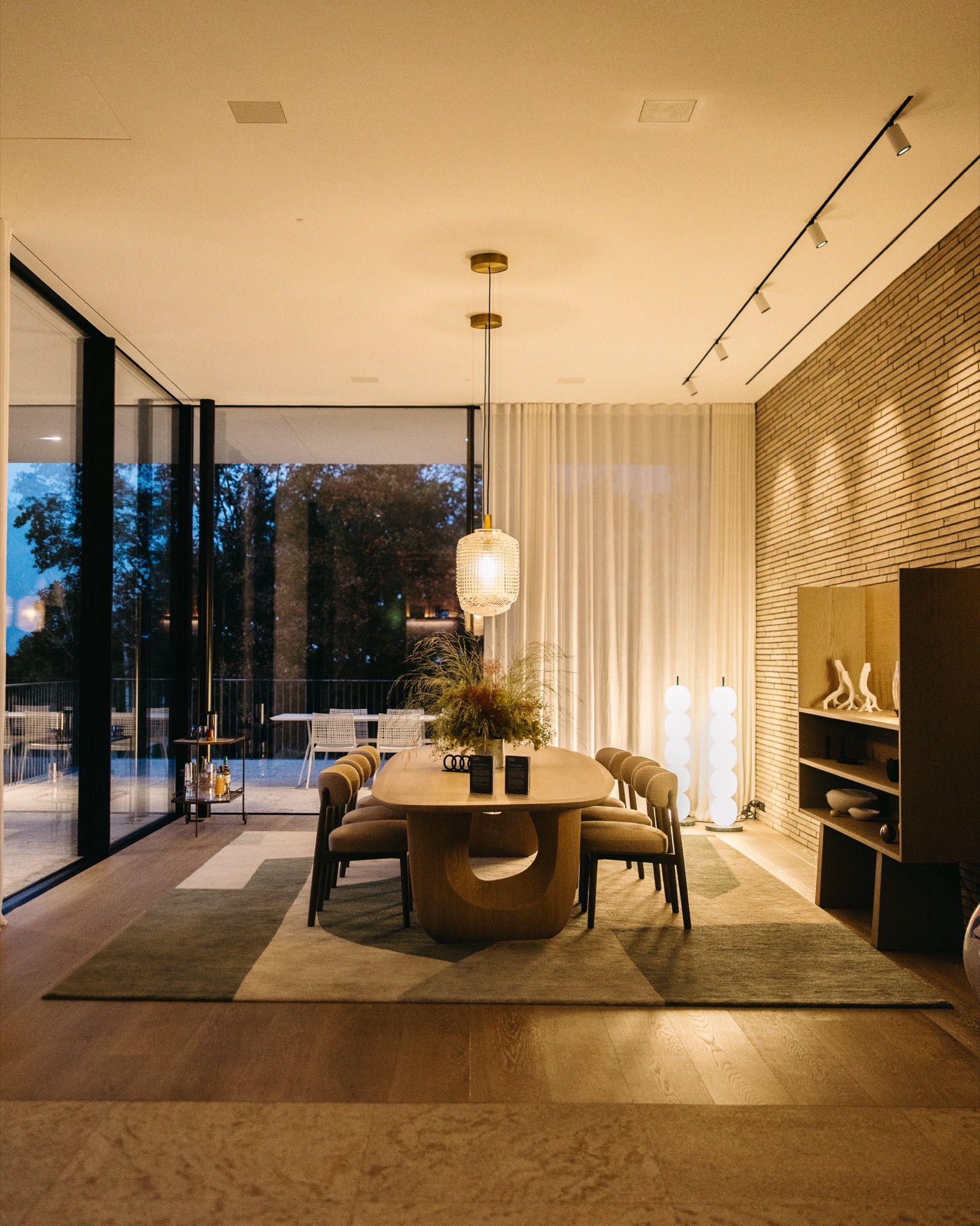 An oval wooden table with six chairs sits on a geometric rug beneath a pendant light, set against floor-to-ceiling windows overlooking an Audi Q9 parked in the dark exterior.